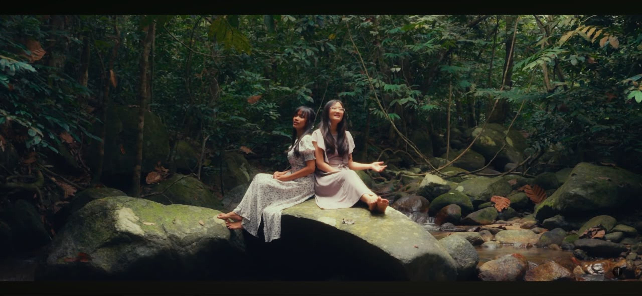 A still from a LoudVoice music video — two singers seated on a rock beside a forest stream